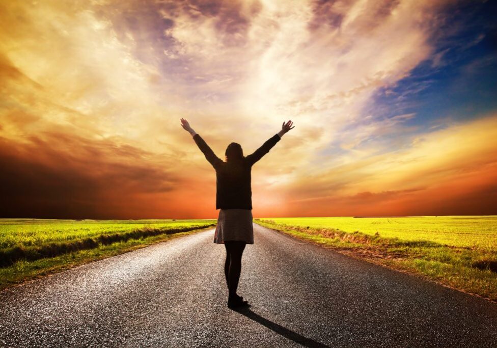 Happy woman standing with hands up on long straight road facing the sun. Sunset sky